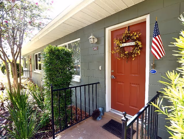 Exterior view of a residential building entrance with a red door decorated with a yellow floral wreath and a small 'Welcome' sign. An American flag is mounted on the wall next to the door. The building has gray siding and white trim, with a black metal railing along the small porch. Green shrubs and plants surround the entrance area.