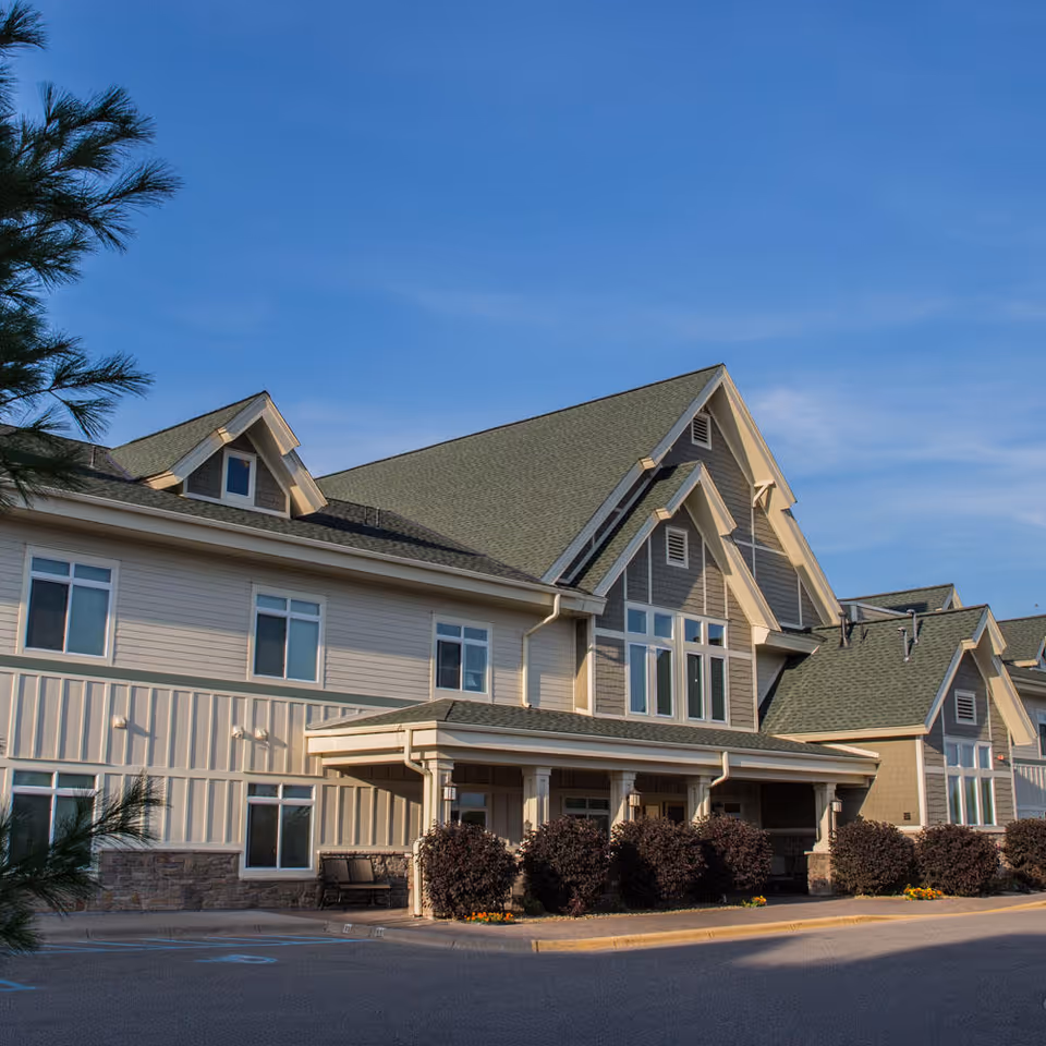 Exterior view of a multi-story senior living facility building with beige and gray siding, multiple windows, and a covered entrance. There are bushes and a tree in the foreground under a clear blue sky.