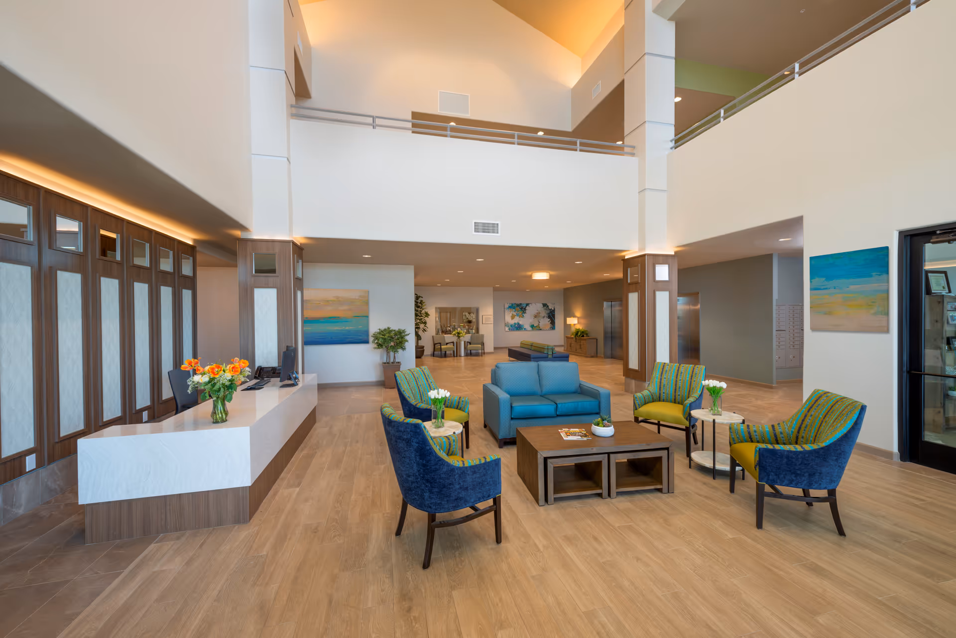 Spacious and modern assisted living facility lobby with high ceilings, a reception desk with flowers, and a seating area featuring blue and green chairs around a wooden coffee table. The space is well-lit with natural and artificial lighting, decorated with paintings and plants.