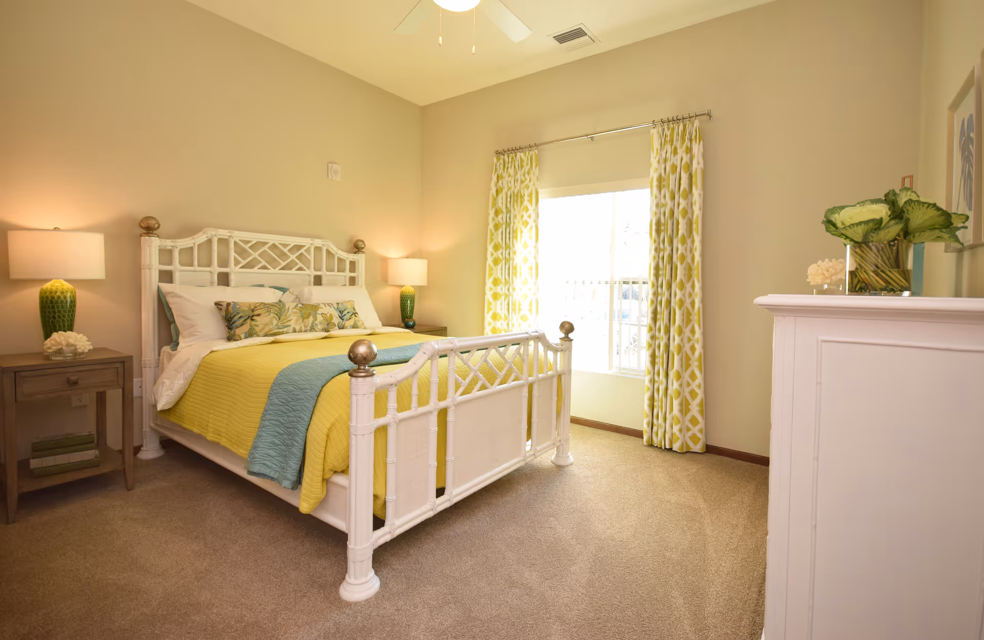 A bright and cozy bedroom with a white wooden bed frame, yellow and blue bedding, two bedside tables with green lamps, patterned yellow curtains on a window, beige walls, and a white dresser with a plant on top.