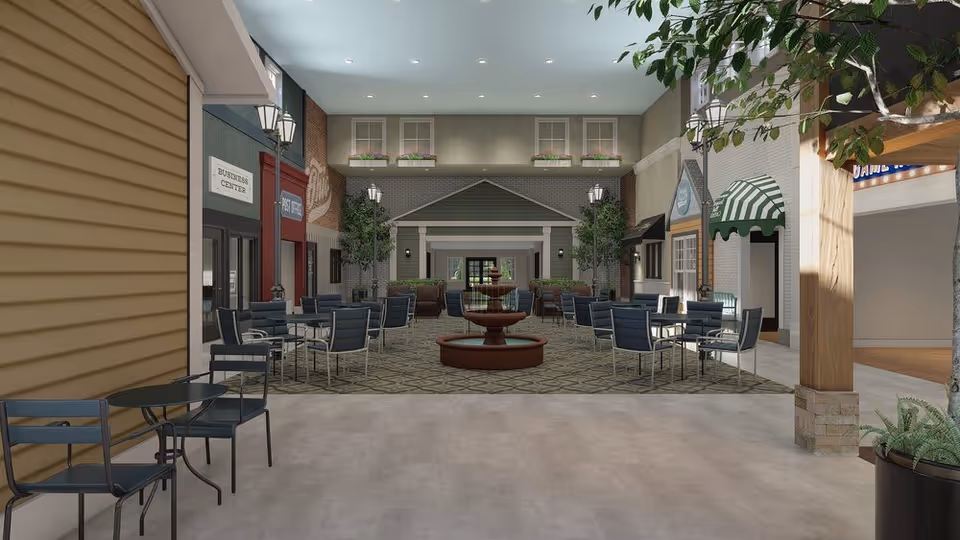 Indoor common area designed like a small village street with multiple seating areas featuring tables and chairs. There is a central fountain surrounded by chairs, decorative street lamps, potted plants, and storefront-style facades labeled Business Center and Post Office. The ceiling is light blue with recessed lighting, and the floor has a patterned carpet in the seating area and smooth flooring in the foreground.