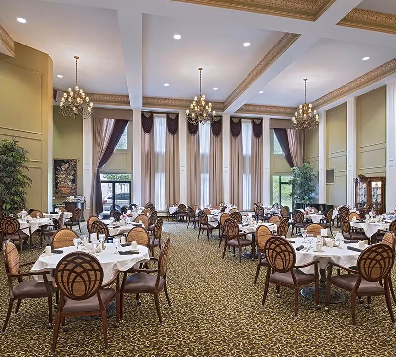 A spacious, elegant dining room with multiple round tables covered with white tablecloths, set with cups, glasses, and napkins. The room features tall windows with beige curtains and dark valances, ornate chandeliers hanging from a high ceiling with decorative molding, and patterned carpet flooring. There are plants and decorative furniture along the walls.