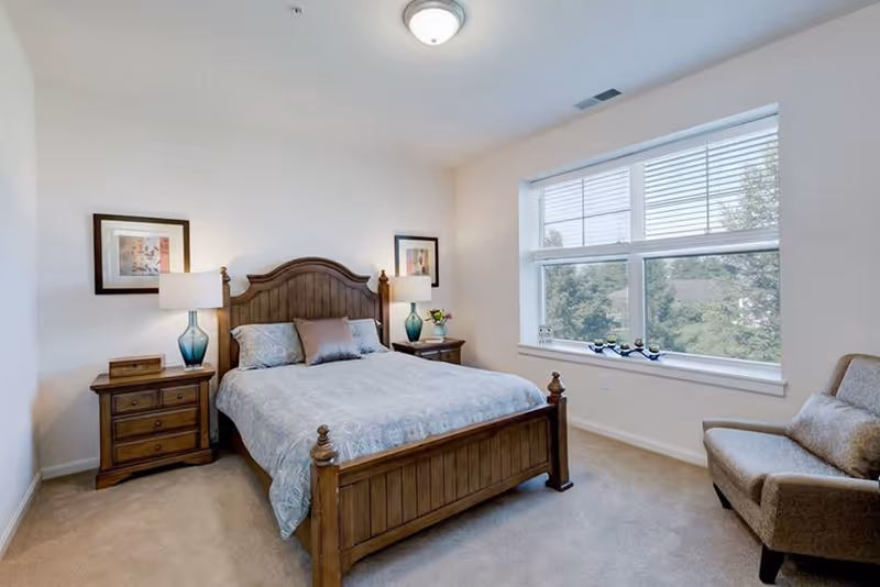 A bright bedroom with a wooden bed frame and matching nightstands on either side. Each nightstand has a blue glass lamp and framed artwork above. There is a large window letting in natural light, and a cushioned armchair with a pillow is positioned near the window.