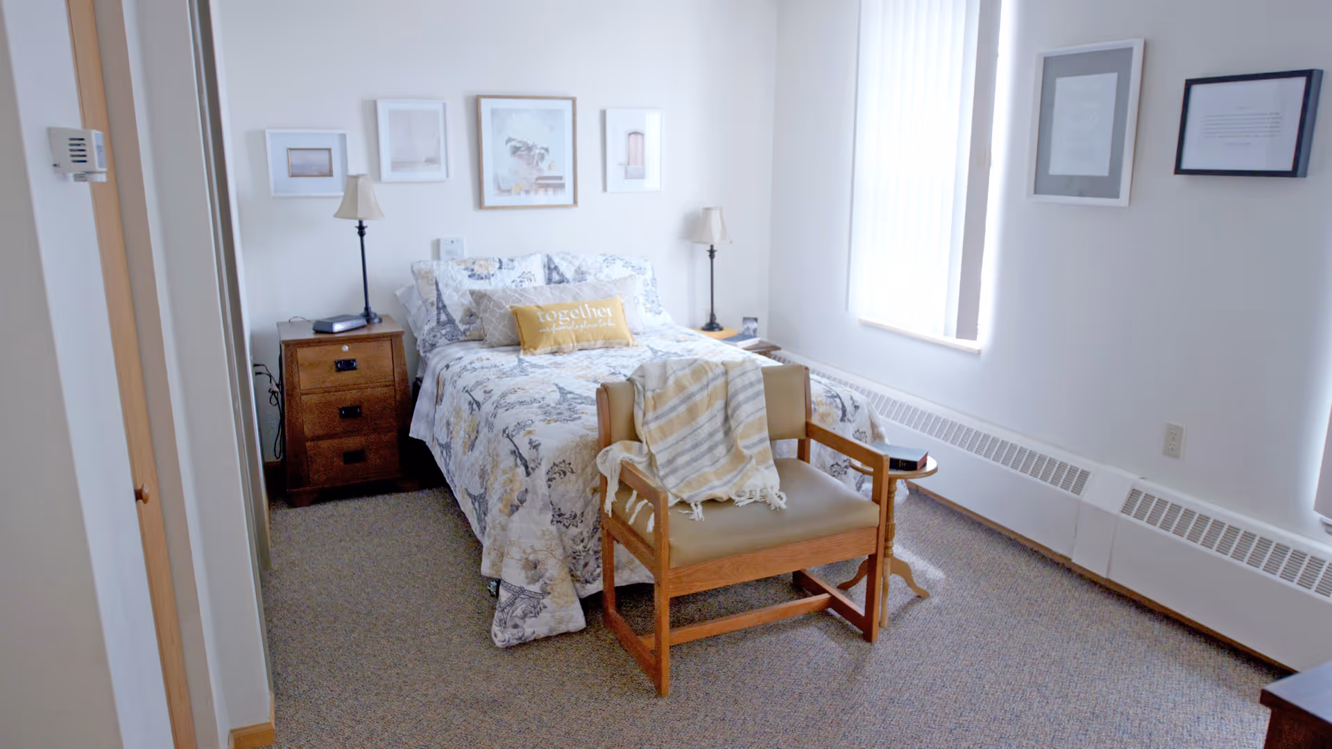 A cozy bedroom with a bed covered in a patterned quilt and a yellow pillow that says 'Together is my favorite place to be.' There are two wooden nightstands with lamps on either side of the bed. A wooden chair with a striped blanket draped over it is placed at the foot of the bed. The room has light-colored walls with framed pictures and a window with vertical blinds letting in natural light.