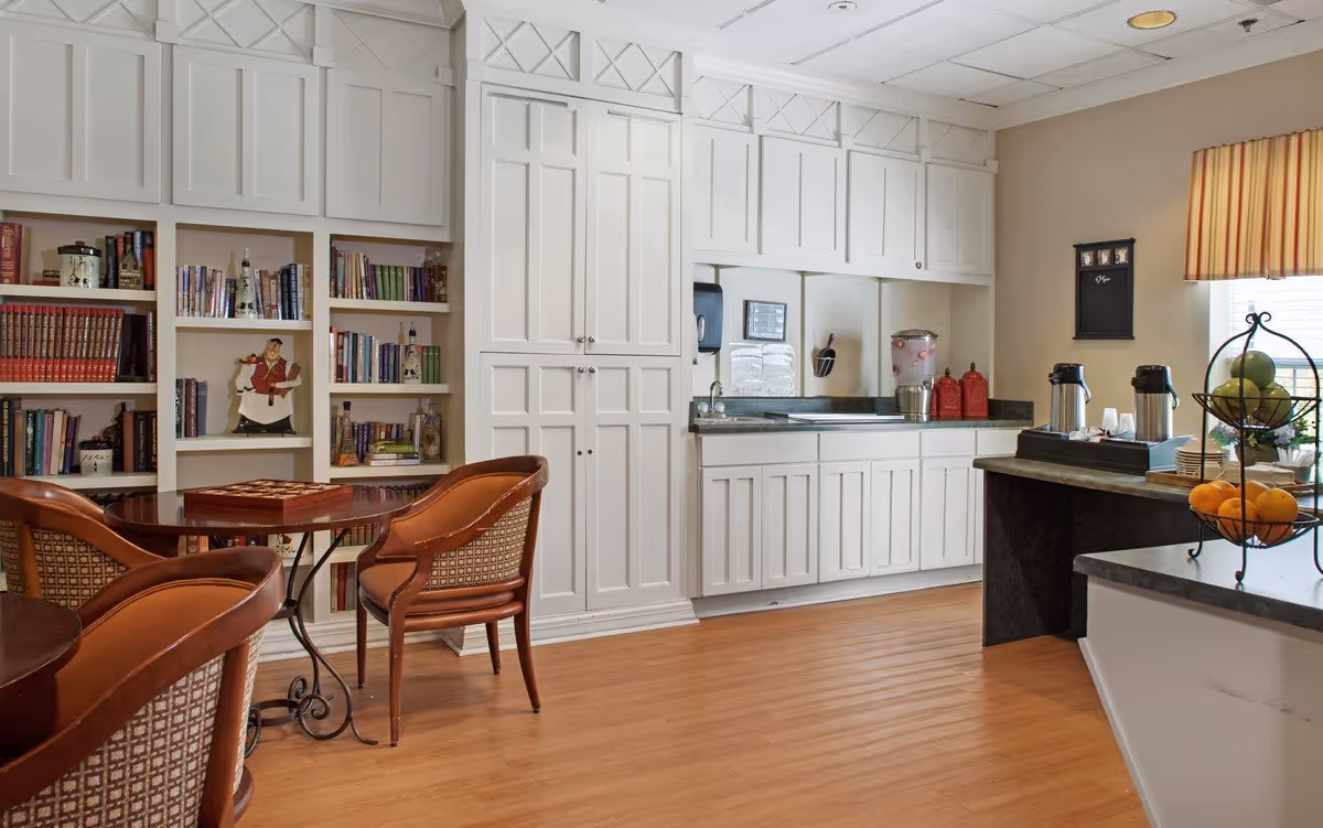 Well-lit communal dining and kitchenette area with bookshelves, a small round table and chairs, and a counter with a beverage station.