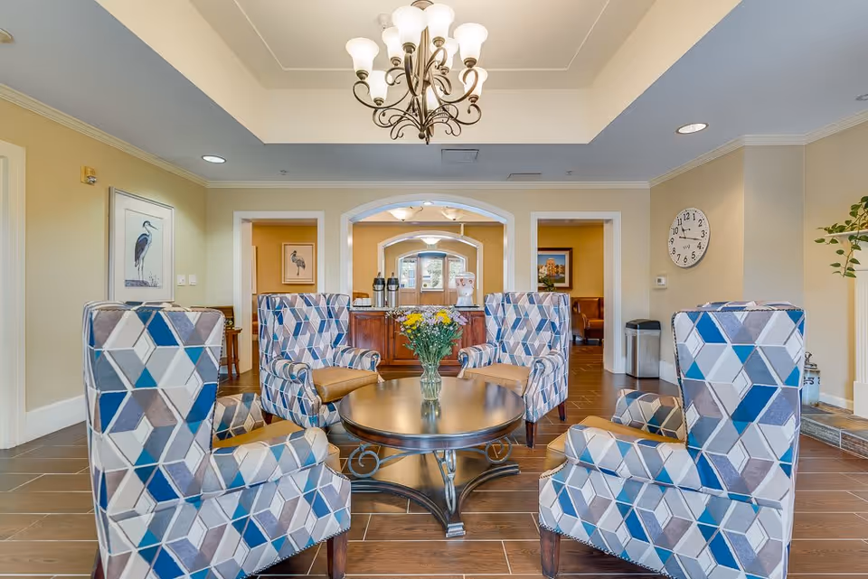 A bright and inviting senior living facility common area with four patterned armchairs arranged around a round wooden coffee table holding a vase of yellow and white flowers. The room features a chandelier, light-colored walls, framed bird artwork, a wall clock, and a wooden sideboard with coffee dispensers in the background.