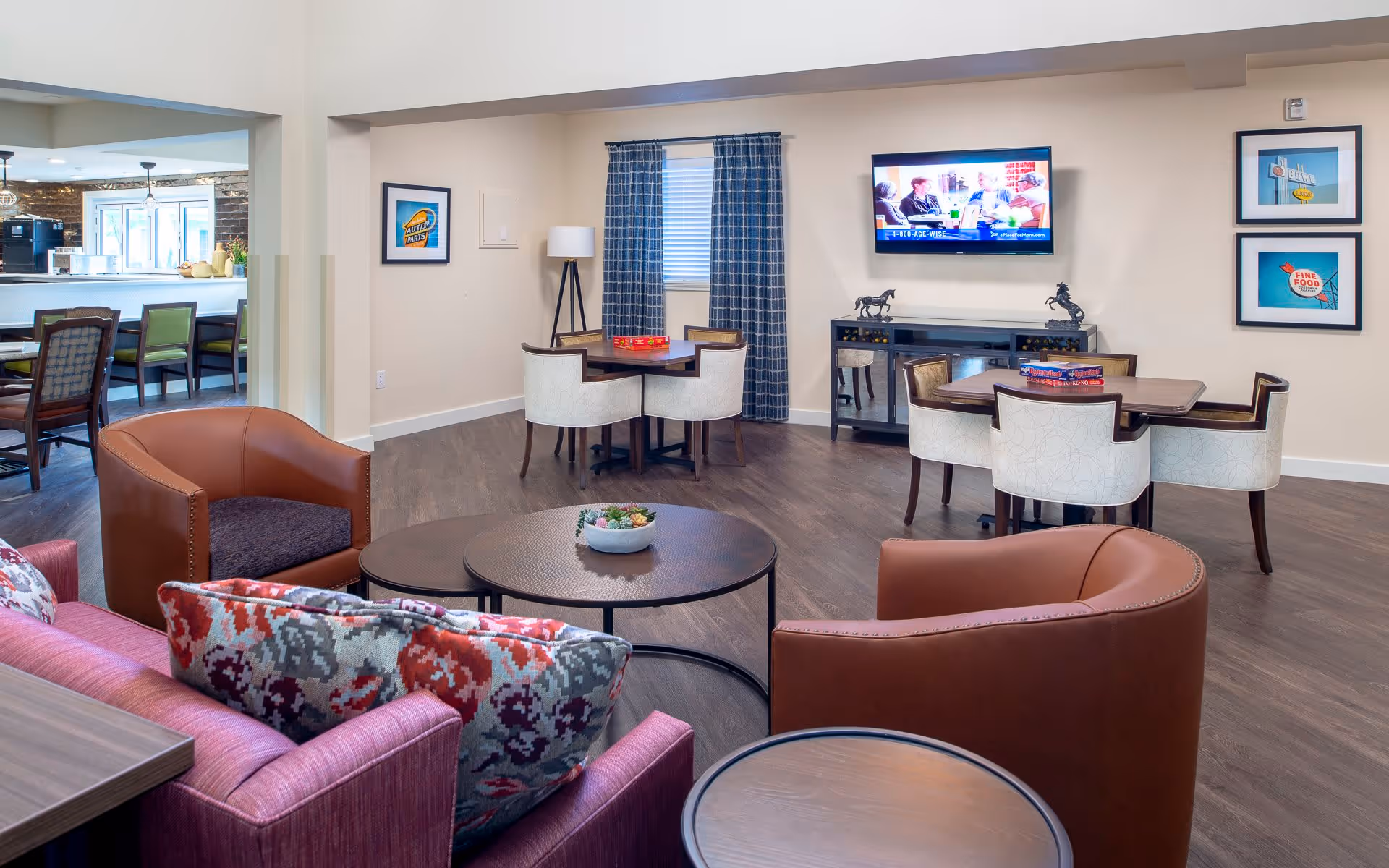 A cozy senior living common area with comfortable seating including a purple sofa with patterned cushions, two brown armchairs, and round coffee tables. In the background, there are two small dining tables with chairs, a wall-mounted TV, framed artwork on the walls, and a window with blue checkered curtains. To the left, there is a view into a kitchen area with bar stools and a counter.