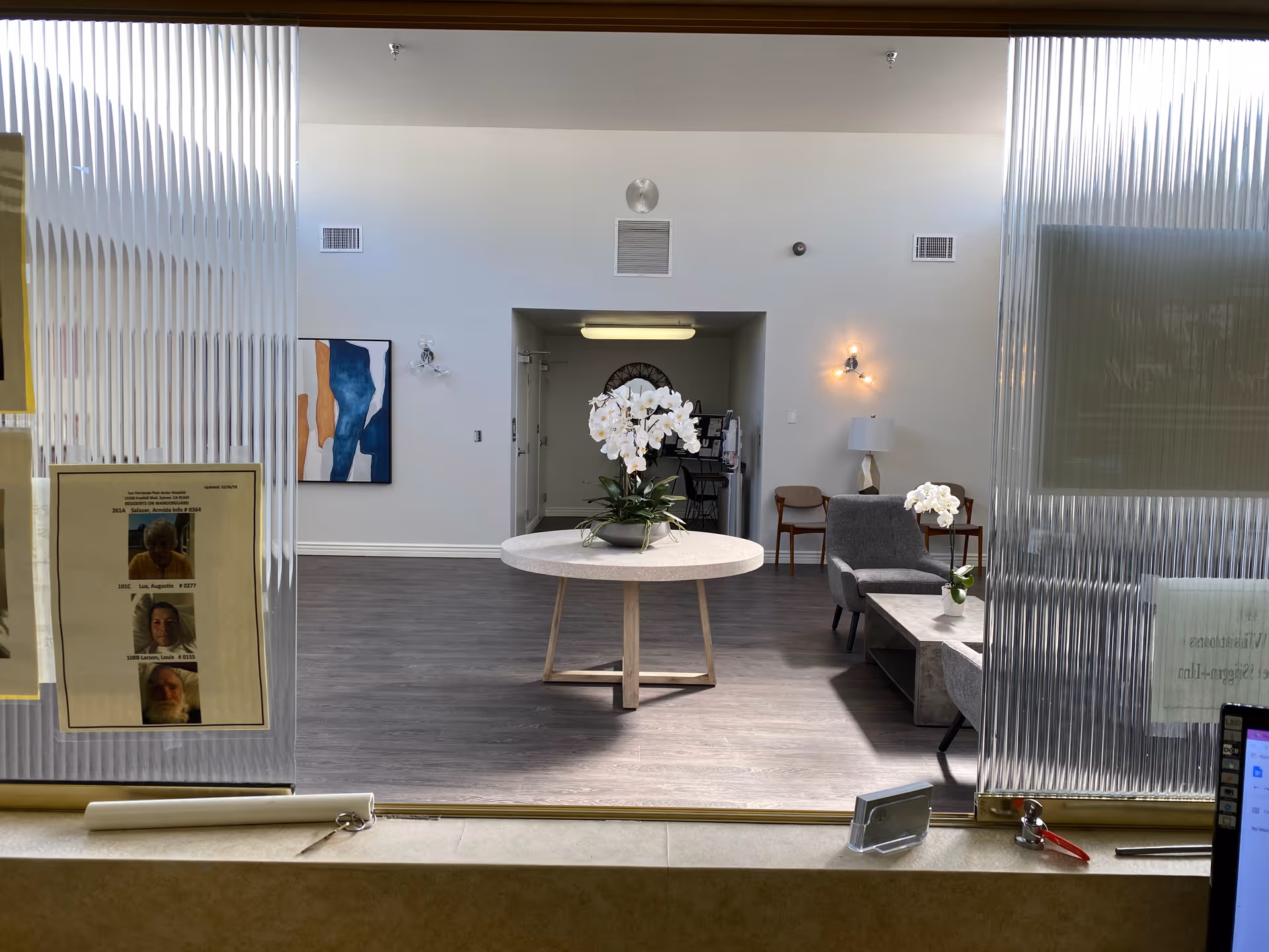 Interior reception/lobby area with a round table holding white orchids, seating chairs and side tables, and artwork on the back wall seen through a service window.