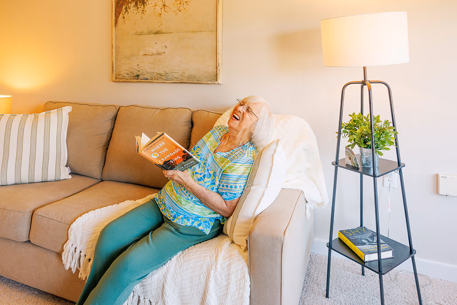 An elderly woman with white hair and glasses is sitting on a beige couch in a cozy living room, laughing while holding an open book titled 'Where the Crawdads Sing.' The couch has a striped pillow and a white throw blanket. Next to the couch is a black metal floor lamp with two shelves, one holding a small green plant and the other holding a book titled 'Shy.' A framed picture of swans on water hangs on the wall behind the couch.