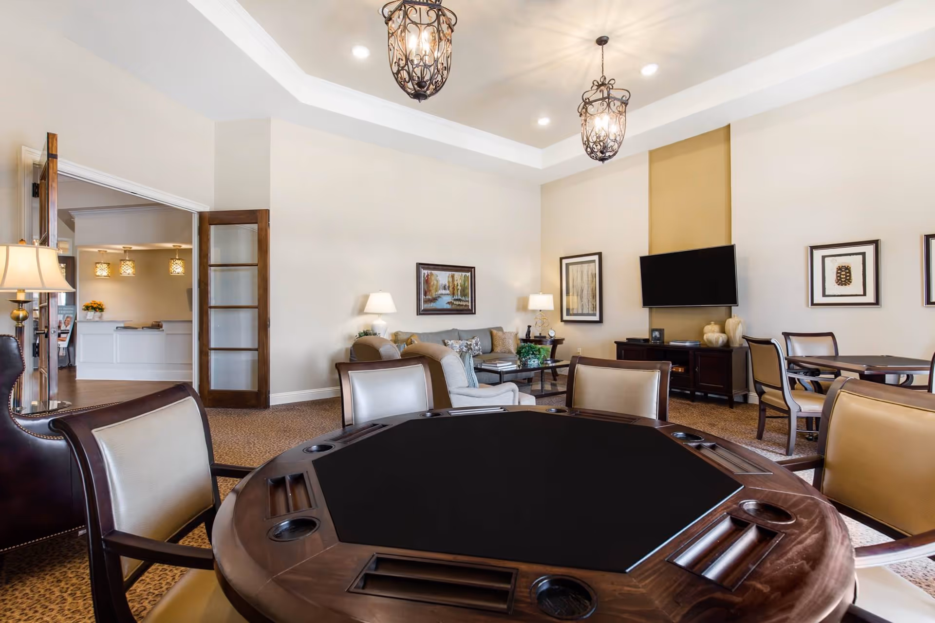 A well-lit senior living common area featuring a round gaming table with chairs in the foreground, comfortable seating with sofas and armchairs, a wall-mounted flat-screen TV, framed artwork on the walls, and decorative hanging light fixtures. The room has a neutral color palette with beige walls and carpeted flooring.