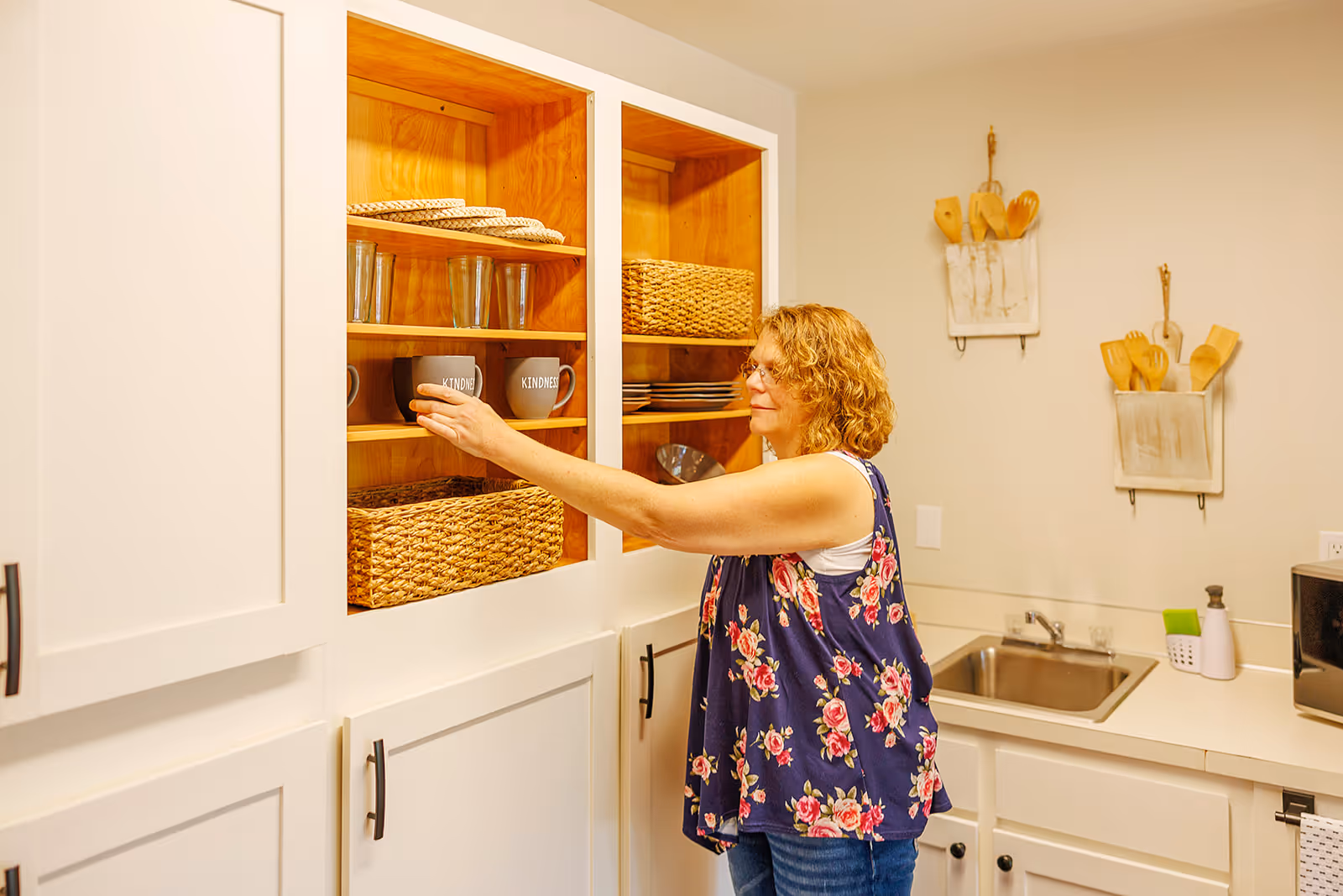 A woman with curly hair wearing a floral sleeveless top is reaching to place or take a mug from a wooden cabinet in a kitchen. The cabinet contains woven baskets, glasses, and mugs. The kitchen has white cabinets, a sink, and wooden utensils hanging on the wall.