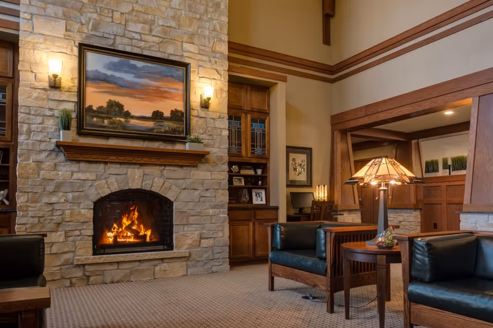 Cozy senior living facility common area with a stone fireplace, a painting above the mantle, two wall sconces, wooden built-in shelves, and comfortable leather armchairs surrounding a wooden side table with a decorative lamp.