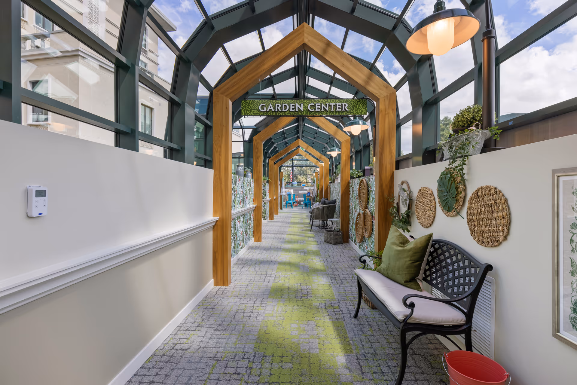 Glass-roofed indoor walkway labeled 'Garden Center' with wooden arch frames, seating bench, decorative plants and patterned carpet.