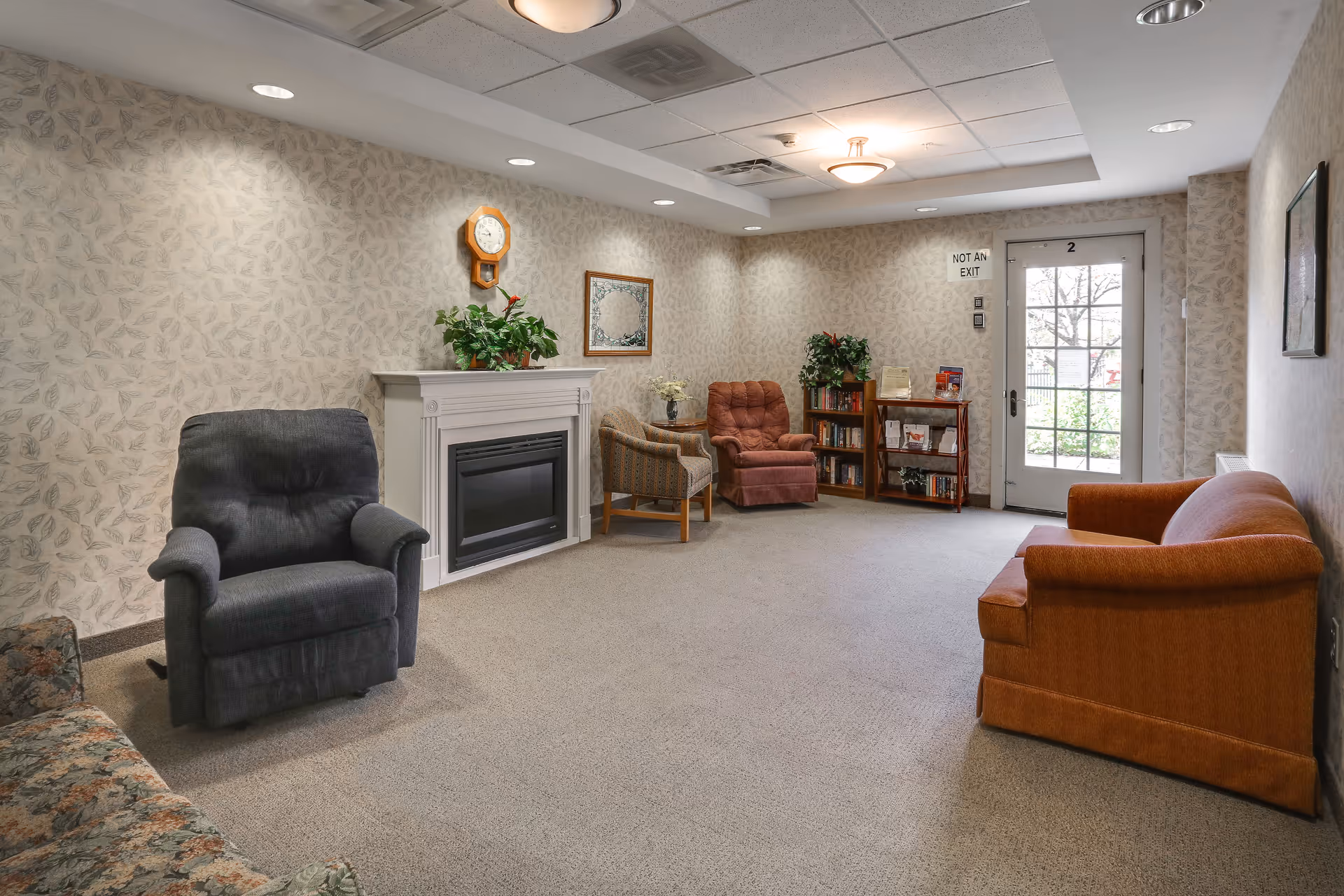 A cozy living room area in a senior living facility with several armchairs and a sofa arranged around a white electric fireplace. The walls have a subtle leaf-patterned wallpaper, and there is a clock and a framed mirror above the fireplace. A small bookshelf and a side table with plants and magazines are near a glass door labeled 'Not an Exit'. The room is carpeted and softly lit with ceiling lights.