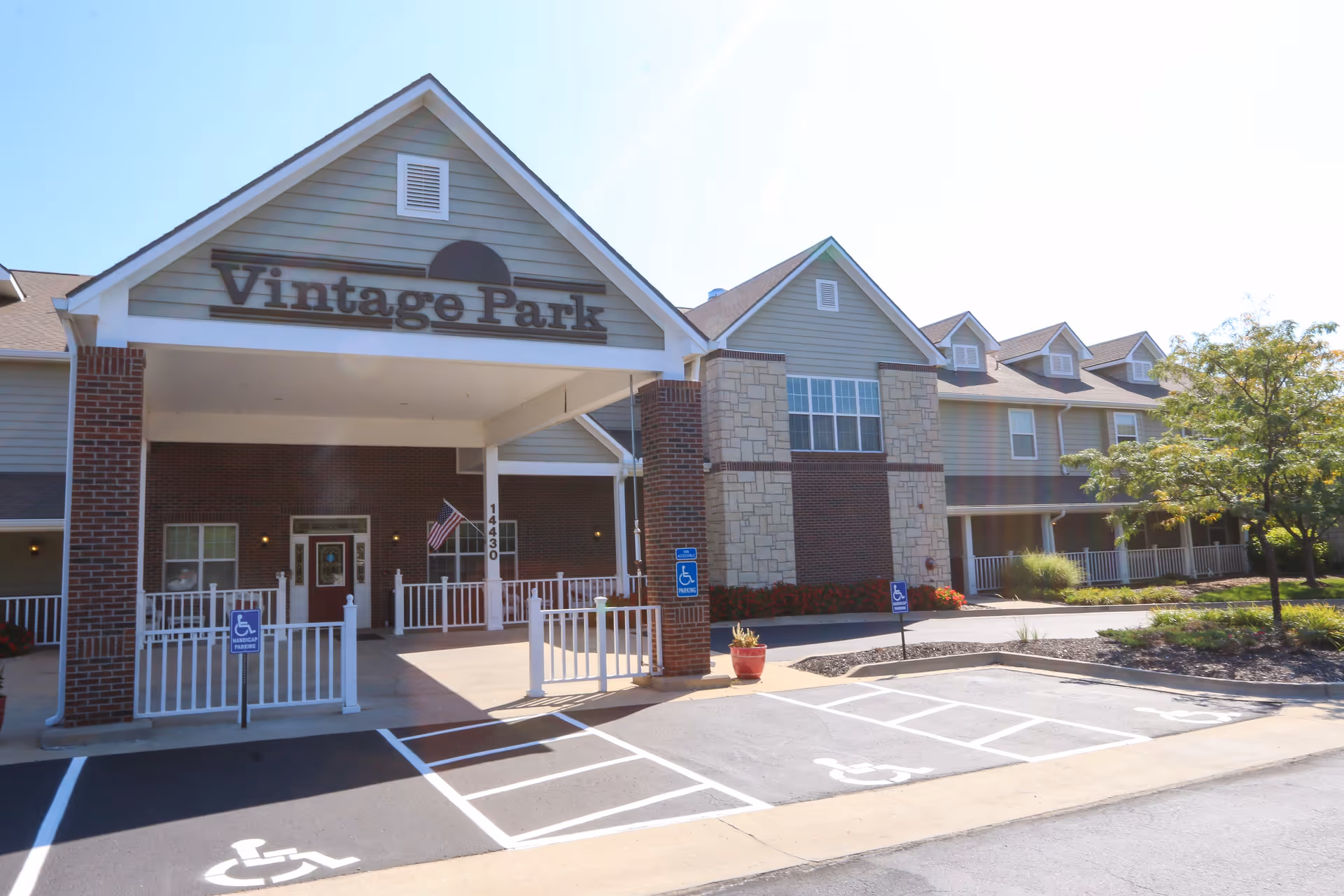 Exterior front entrance of Vintage Park at Stanley senior living facility with a covered drop-off area, brick and stone facade, handicap parking spaces, and a small landscaped area with a tree.