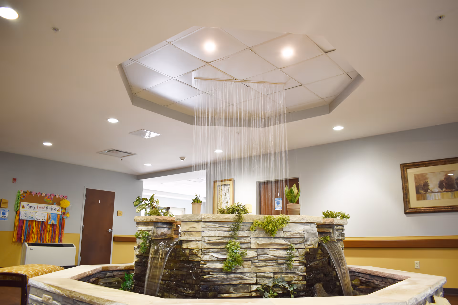 Indoor water fountain feature with cascading water streams in a senior living facility lobby area, surrounded by stone walls and decorated with green plants. The ceiling above has recessed lighting and a geometric design. The background shows doors, a bulletin board, and framed artwork on the walls.