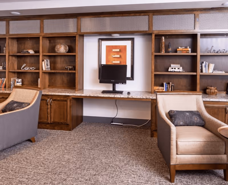A cozy interior room with built-in wooden bookshelves filled with books and decorative items. There is a granite countertop with a computer monitor and keyboard placed on it. Two cushioned armchairs with pillows are positioned in front of the shelves. The carpeted floor and warm wood tones create a comfortable atmosphere.