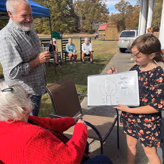 A young girl holding a whiteboard with a drawing and the words 'leaves can fall down' written on it, standing outdoors near a seated elderly woman in a red sweater. A man with a beard and plaid shirt is standing nearby smiling. In the background, two elderly people are seated on chairs on the grass, with trees and a small shed visible.