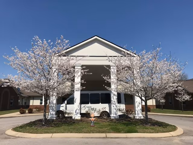 Front exterior view of Grace Manor Assisted Living building with a covered entrance supported by white columns, two blooming trees on either side, and a white van parked under the entrance.