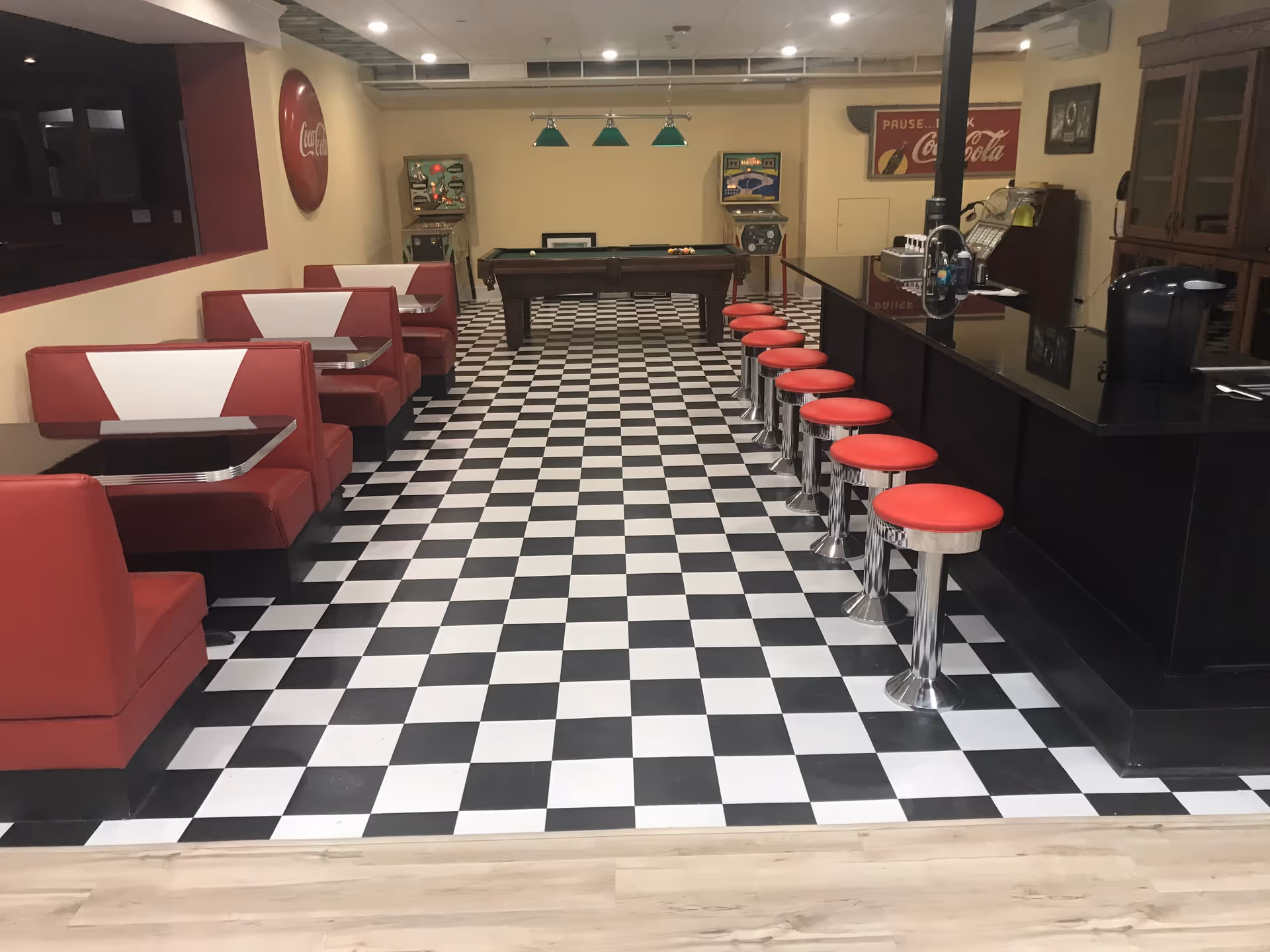 Interior view of a retro-style diner area with black and white checkered floor, red and white booth seating on the left, a black counter with red cushioned stools on the right, and a pool table in the background under green hanging lights. Vintage Coca-Cola signs and pinball machines are visible on the walls.