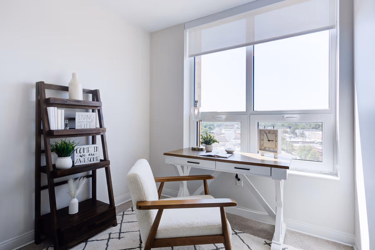 A bright room with a wooden desk positioned in front of a large window. On the desk are a small plant, a clock, a notebook, and a glass ornament. To the left of the desk is a dark wooden ladder shelf holding decorative items including books, a plant, and framed signs. A cushioned chair with wooden armrests is placed in front of the desk on a patterned rug.