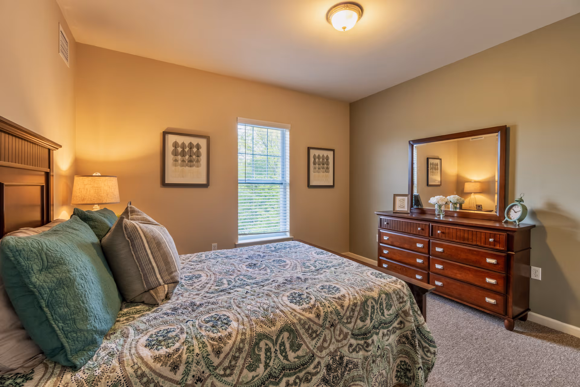 A cozy bedroom with a large bed covered in a patterned quilt and multiple pillows. There is a wooden headboard, a bedside lamp, two framed pictures on the wall, a window with blinds letting in natural light, and a wooden dresser with a large mirror, decorative items, and a clock.