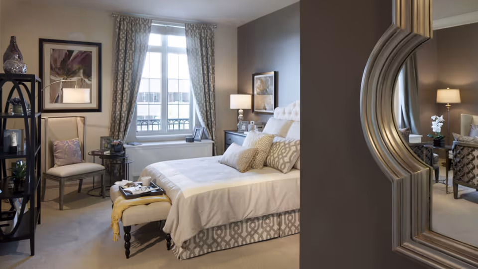A well-decorated bedroom in a senior living facility with a large bed featuring multiple pillows and a white comforter. There is a window with patterned curtains letting in natural light, a chair with a cushion, a small round side table, a shelving unit with decorative items, and a large ornate mirror reflecting part of the room. The room has soft lighting from table lamps and framed artwork on the walls.