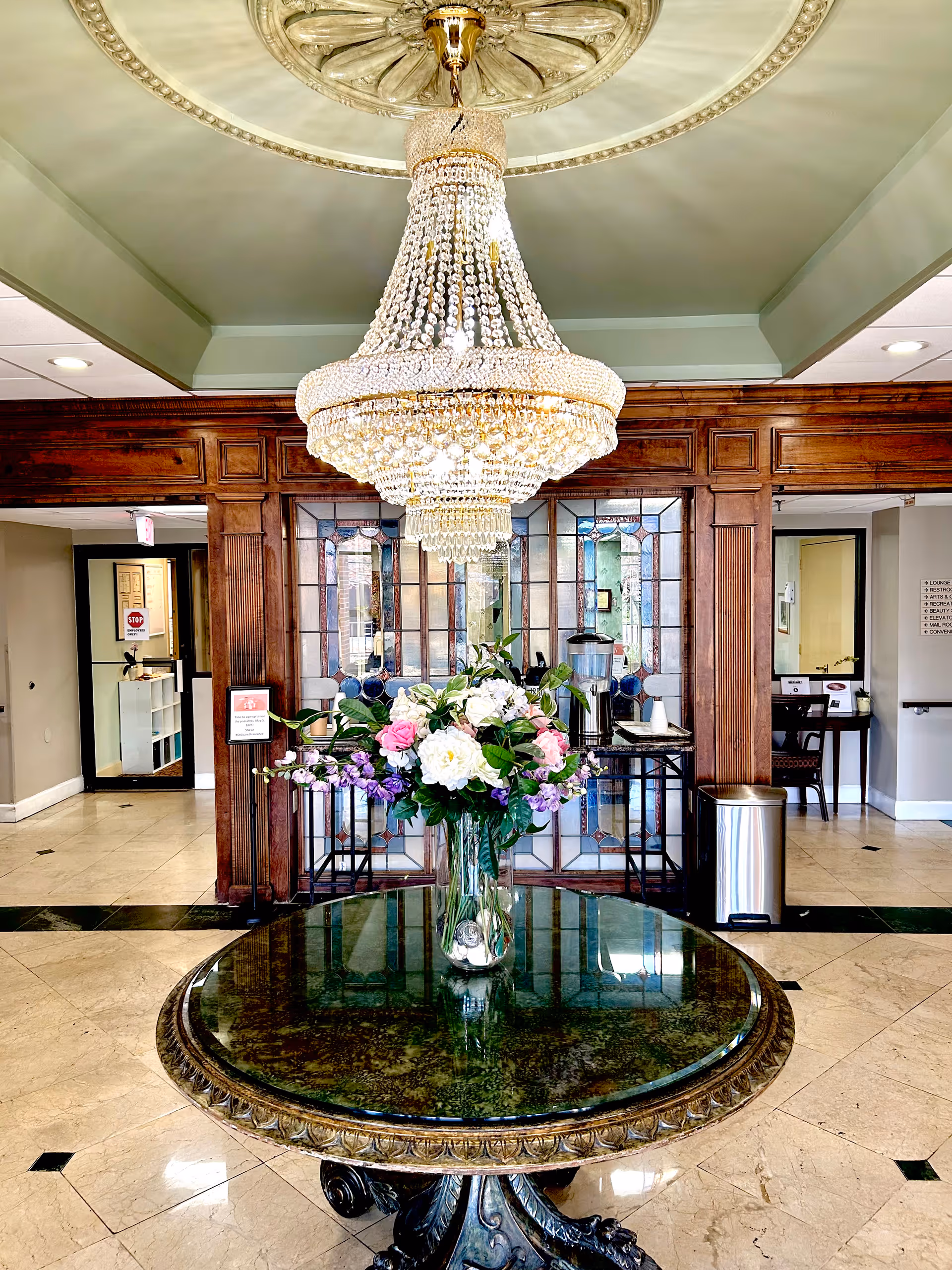 Interior view of a senior living facility lobby featuring a large ornate chandelier hanging from the ceiling above a round table with a glass top. On the table is a vase filled with a colorful bouquet of flowers. The background shows wooden framed glass panels and doorways leading to other rooms.
