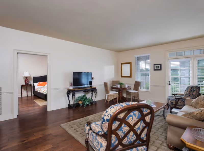 Spacious furnished living room with patterned armchairs, a sofa, TV on a console, a small dining table, and a doorway into a bedroom.