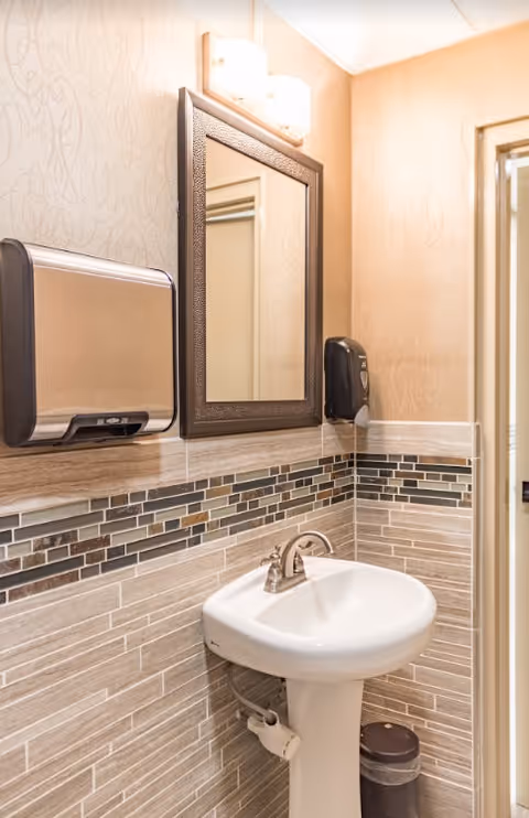 A small bathroom with a white pedestal sink, a rectangular mirror with a dark frame above it, a wall-mounted paper towel dispenser on the left, a soap dispenser on the right, and a trash can below the sink. The walls have beige wallpaper above a tiled wainscoting with a mosaic tile border.