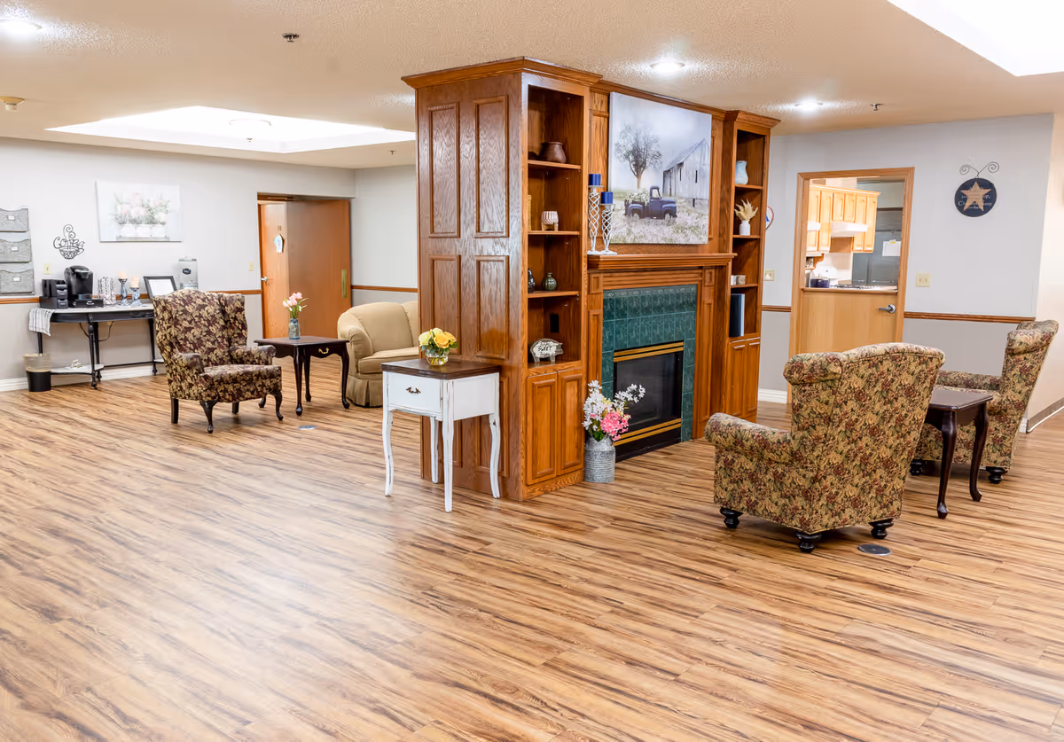 A bright senior living lounge with wood floors, upholstered chairs, side tables, and a central wooden fireplace shelving unit.