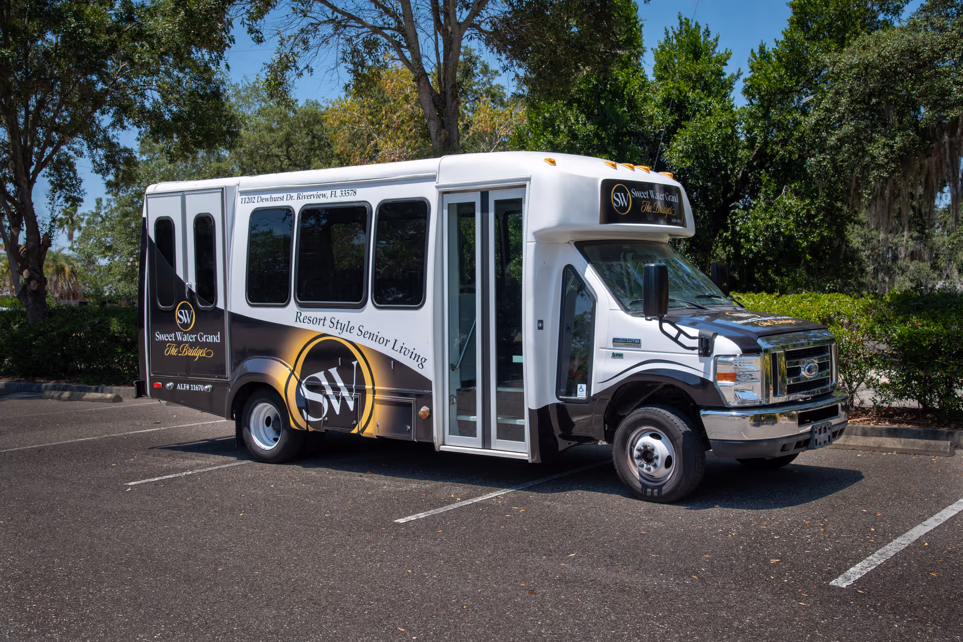 A white and black shuttle bus parked in an outdoor parking lot surrounded by trees. The bus has signage for Sweet Water Grand The Bridges Assisted Living, including the address 11102 Dewhurst Dr, Riverview, FL 33578 and the slogan Resort Style Senior Living.