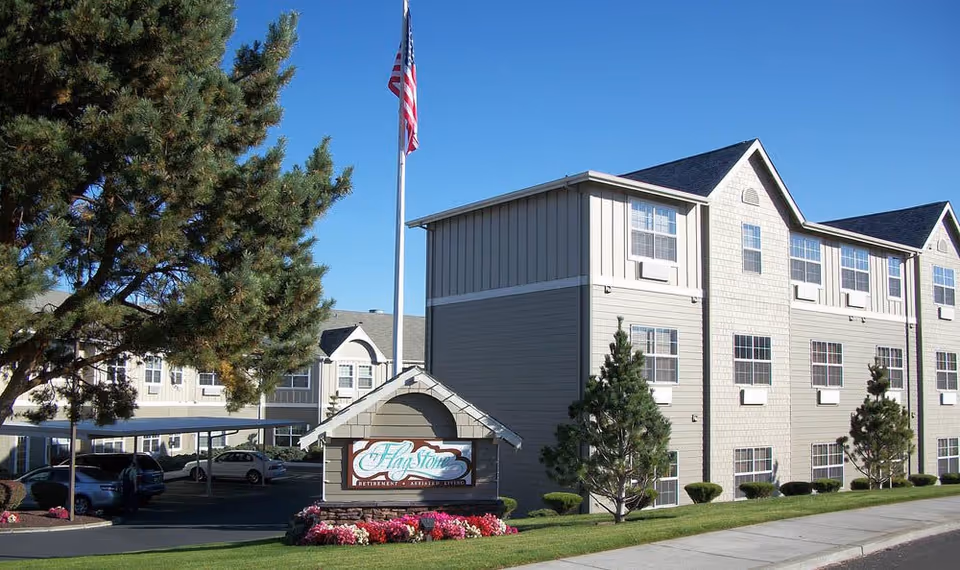 Exterior view of Flagstone Senior Living facility showing a multi-story building with multiple windows, a flagpole with an American flag, a sign with the facility name surrounded by flowers, and a parking area with cars under a covered structure.