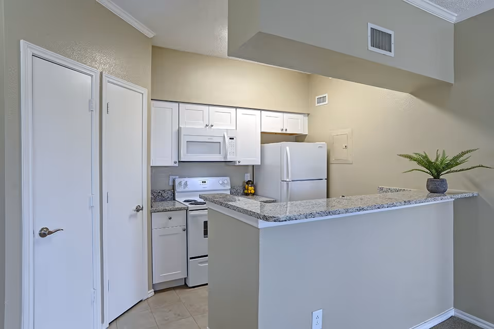Small kitchen area with white cabinets, a white microwave, stove, and refrigerator. There is a granite countertop with a small potted plant on the right side. Two closed white doors are visible on the left side of the image. The walls are painted beige.