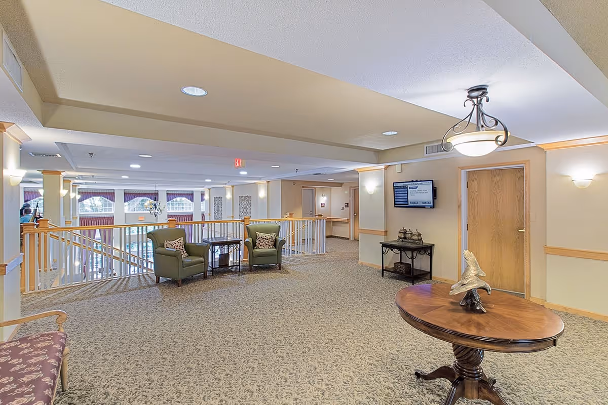 A spacious, well-lit common area in an assisted living facility featuring carpeted floors, a round wooden table with a decorative sculpture, two green armchairs with a small table between them, a wall-mounted TV, and a wooden door. The area overlooks a lower floor with large windows and purple curtains.