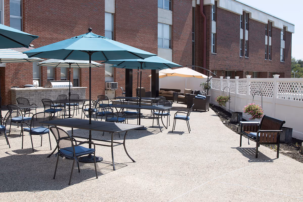Outdoor patio area with multiple tables and chairs, some shaded by large blue umbrellas. There is a brick building in the background and a white fence with hanging flower baskets along the right side. A seating area with cushioned chairs and a beige umbrella is visible in the back corner.