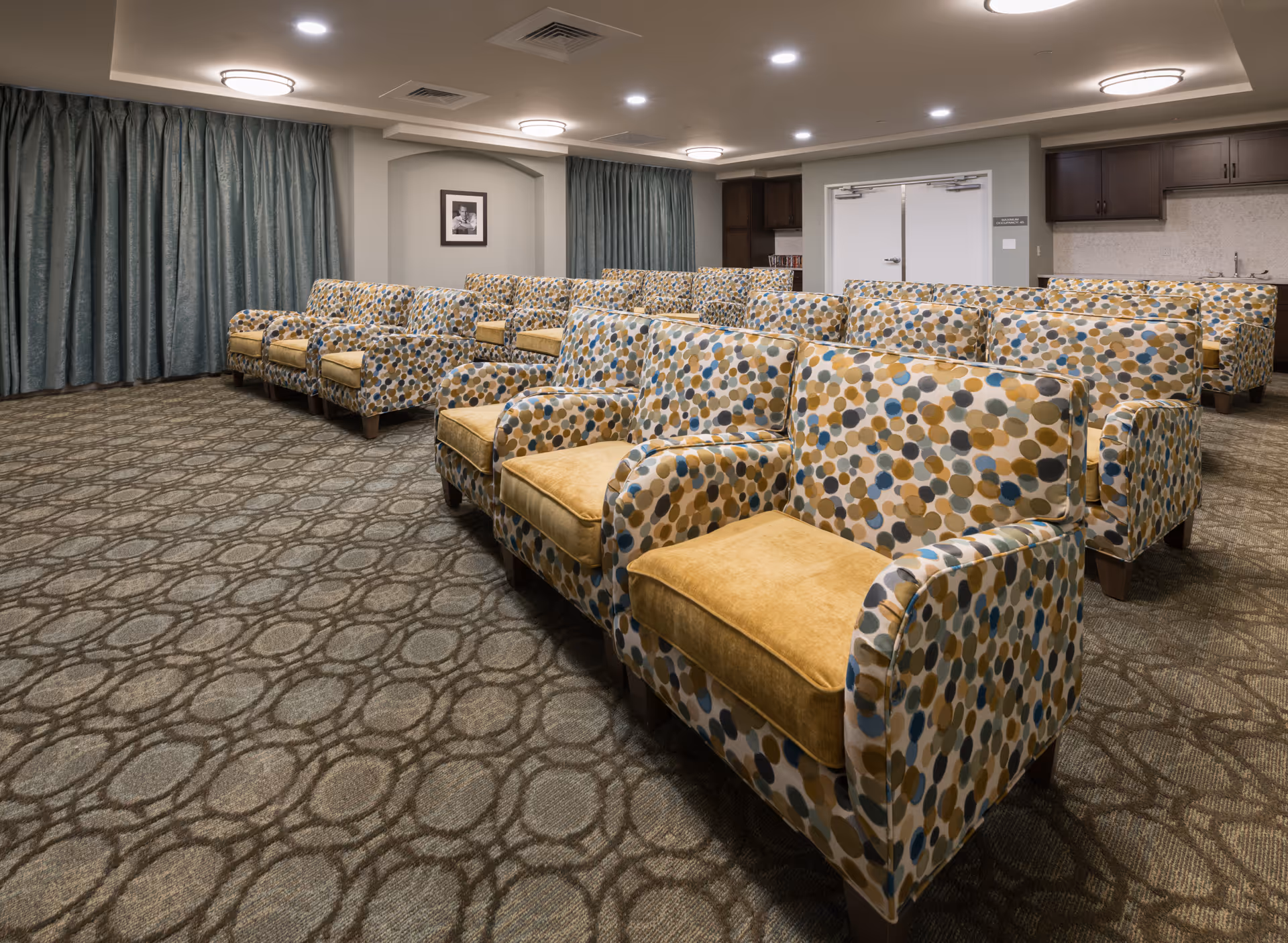 A room with multiple rows of patterned armchairs featuring yellow cushions, arranged facing forward. The room has patterned carpet flooring, grayish-blue curtains covering the windows, and a kitchenette area with dark cabinets and a sink in the back right corner. The walls are light-colored with ceiling lights providing illumination.