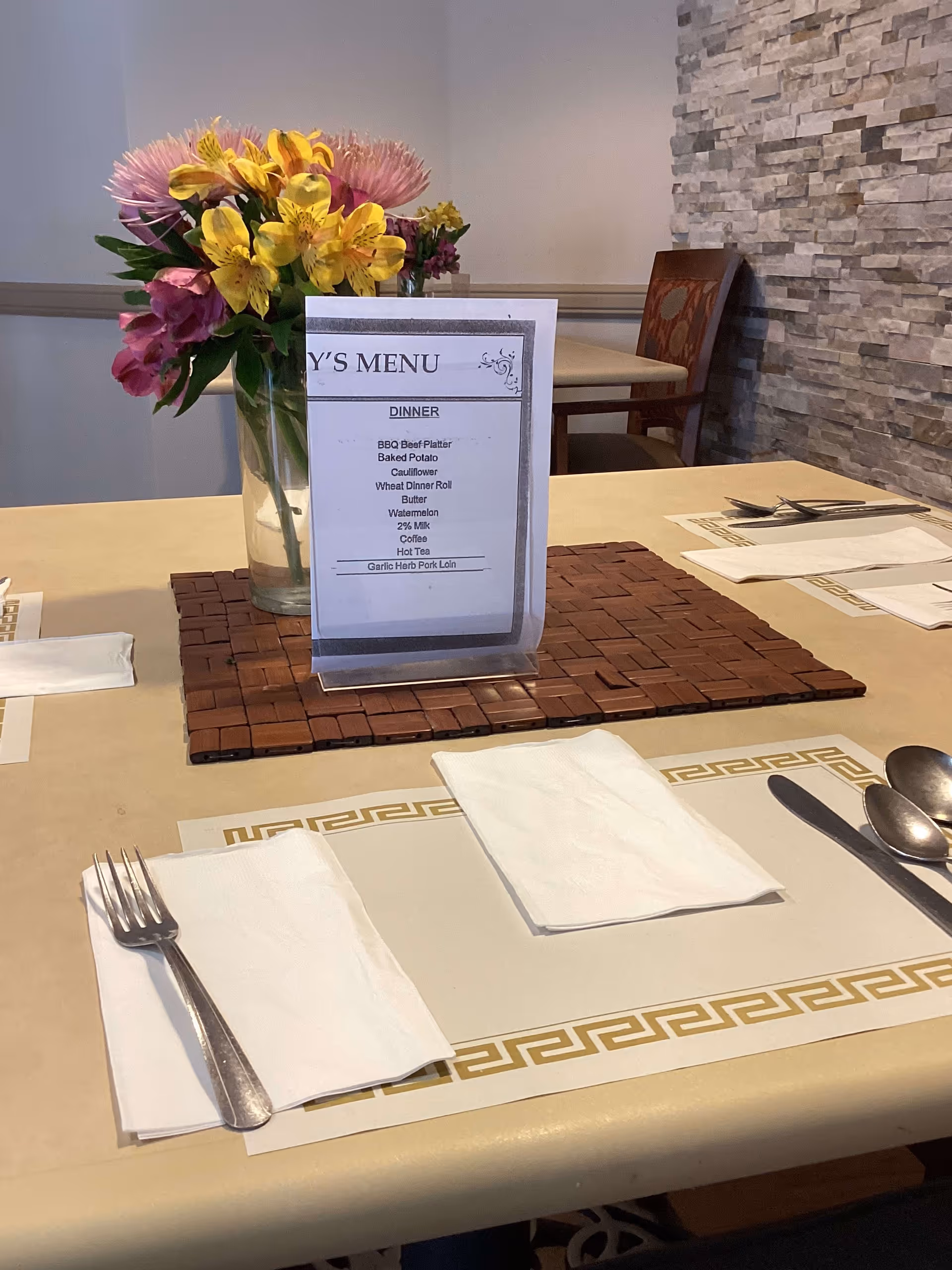 A dining table set with placemats, napkins, forks, spoons, and knives. In the center of the table is a vase with colorful flowers and a menu card listing dinner items including BBQ Beef Platter, Baked Potato, Cauliflower, Wheat Dinner Roll, Butter, Watermelon, 2% Milk, Coffee, Hot Tea, and Garlic Herb Pork Loin. The background shows a stone accent wall and a wooden chair.