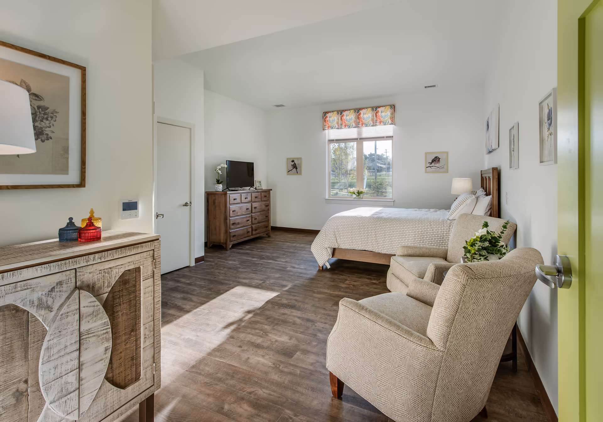 Sunlit furnished bedroom with a bed, wooden dresser and TV, and two upholstered armchairs near a window.