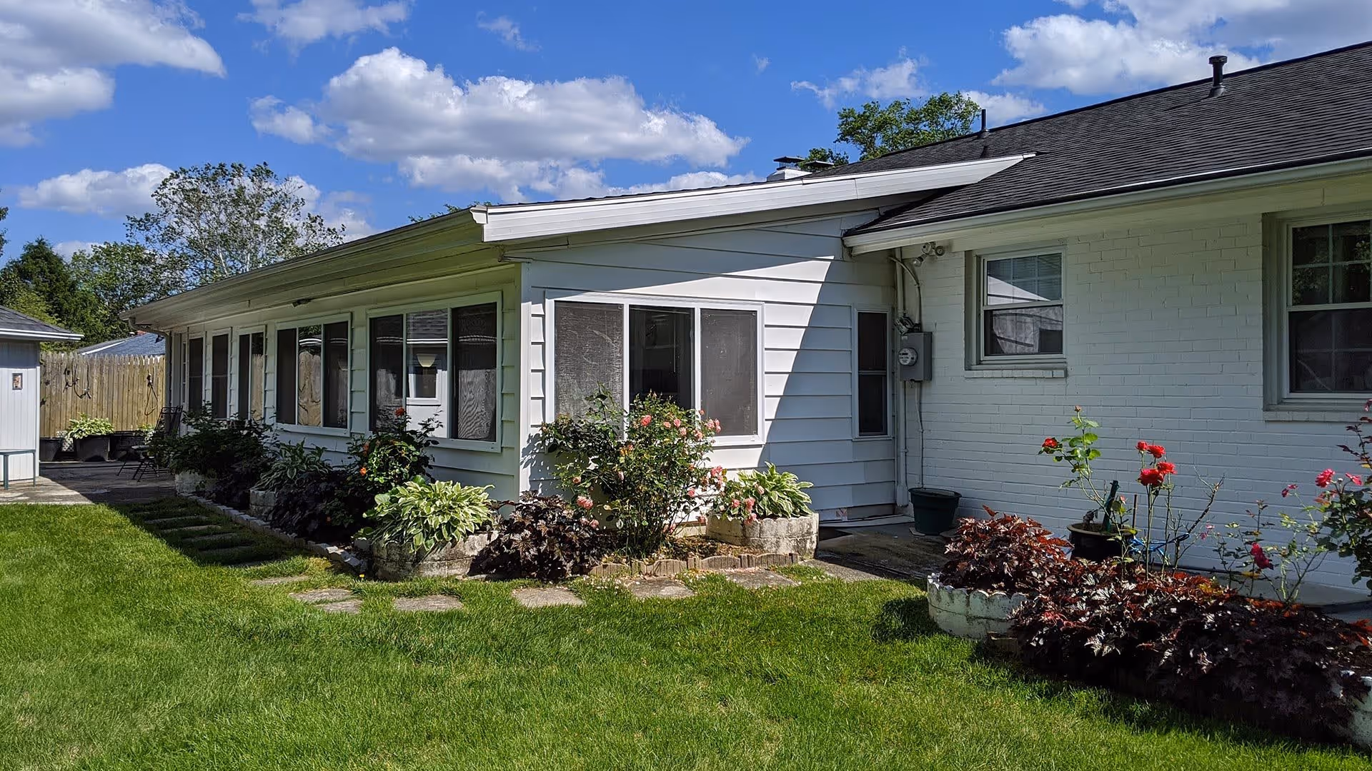Exterior view of a single-story white building with a well-maintained lawn and flower beds featuring various plants and flowers under a blue sky with scattered clouds.