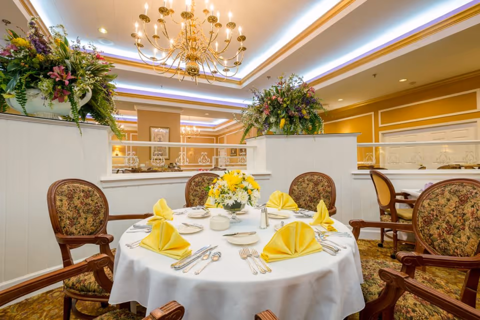 A round dining table set with white tablecloth, yellow folded napkins, silverware, and a floral centerpiece in a well-lit dining room with ornate chairs, floral arrangements on white partitions, and a large chandelier overhead.