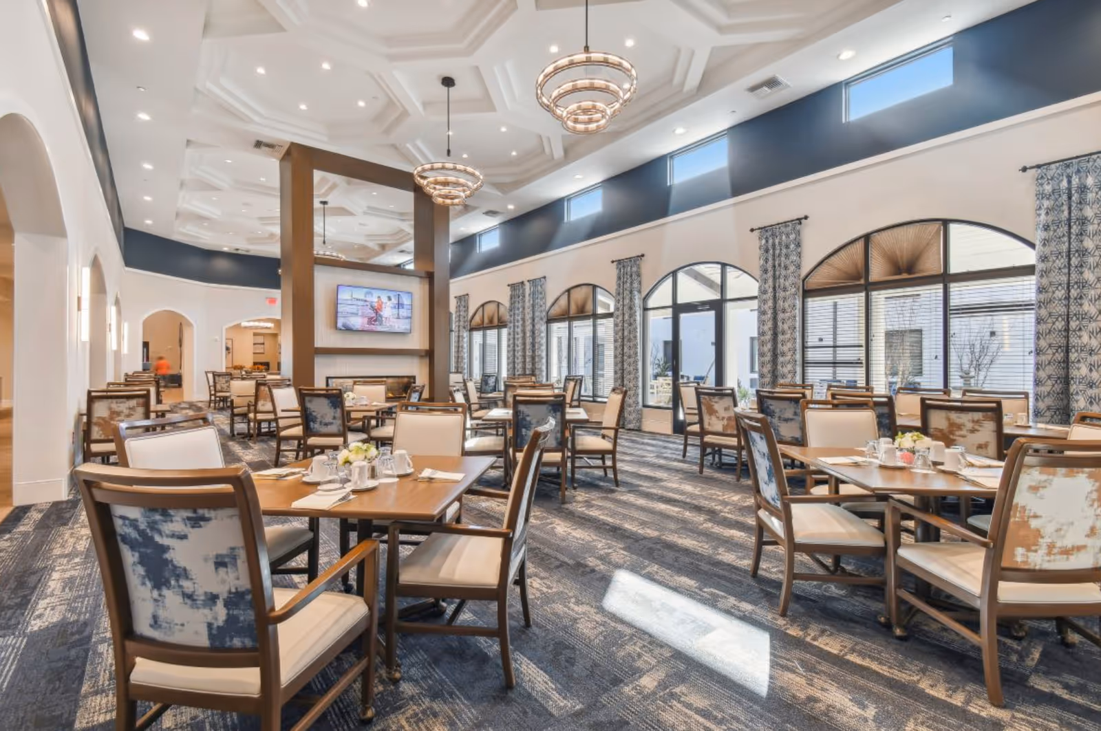 A spacious and well-lit dining room with multiple wooden tables and cushioned chairs arranged neatly. The room features large arched windows with patterned curtains, a high ceiling with decorative molding, and modern circular chandeliers. A flat-screen TV is mounted on a wooden partition in the center of the room.