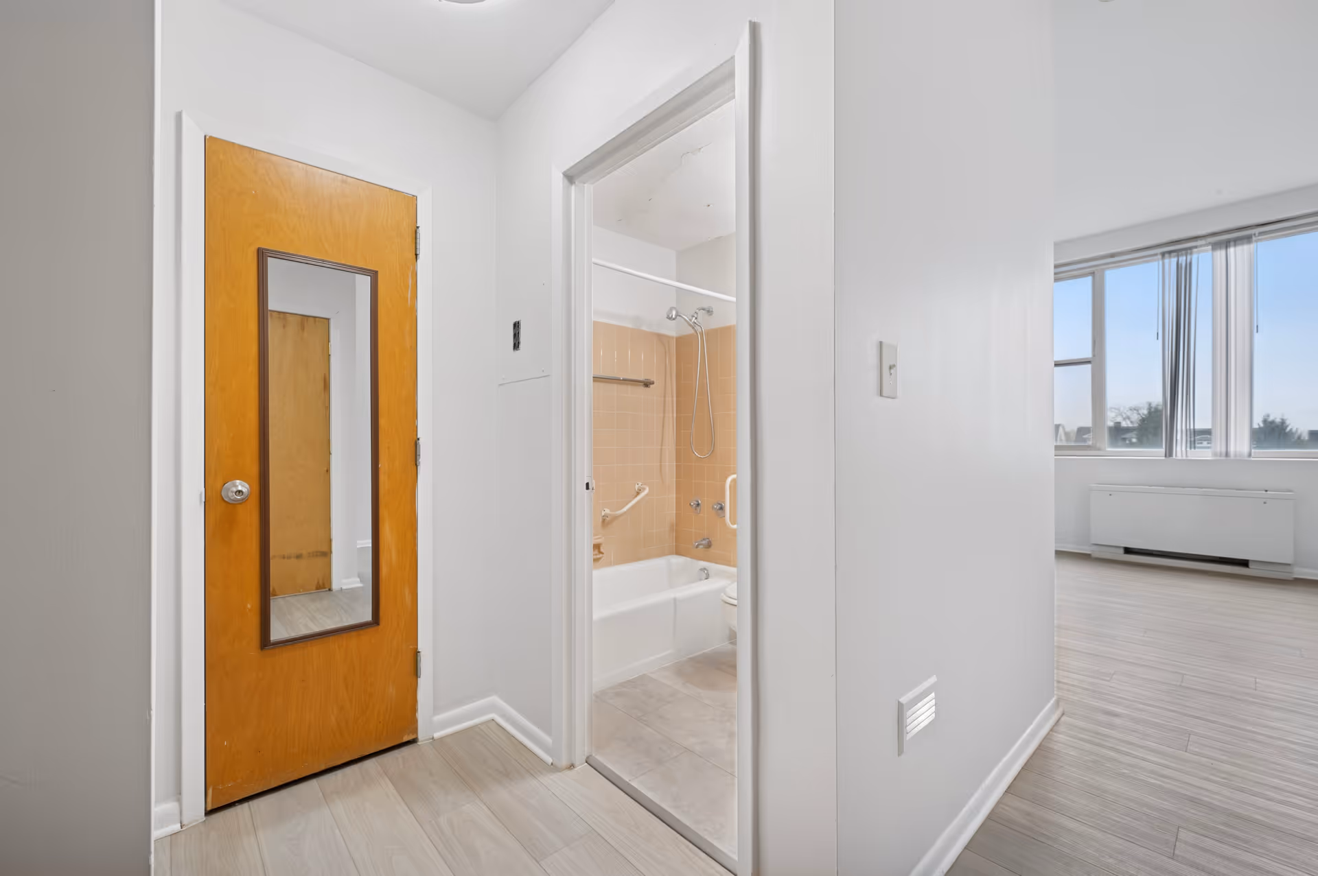 Interior view of an apartment hallway showing a wooden door with a mirror, an open doorway leading to a bathroom with a bathtub and beige tiles, and a bright living area with large windows and light-colored flooring.