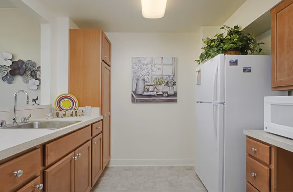 A kitchen with wooden cabinets, a white refrigerator with plants on top, a microwave on the counter, a sink with a faucet, and decorative items including a colorful plate and a painting of flowers on the wall.