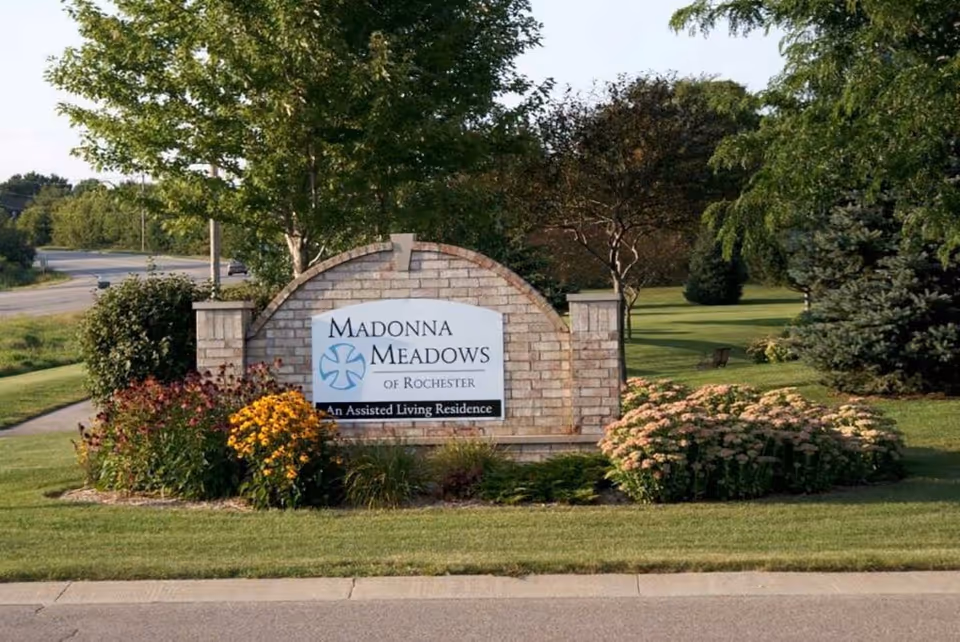 Brick entrance sign reading 'Madonna Meadows of Rochester' surrounded by flowers, trees, and a manicured lawn.