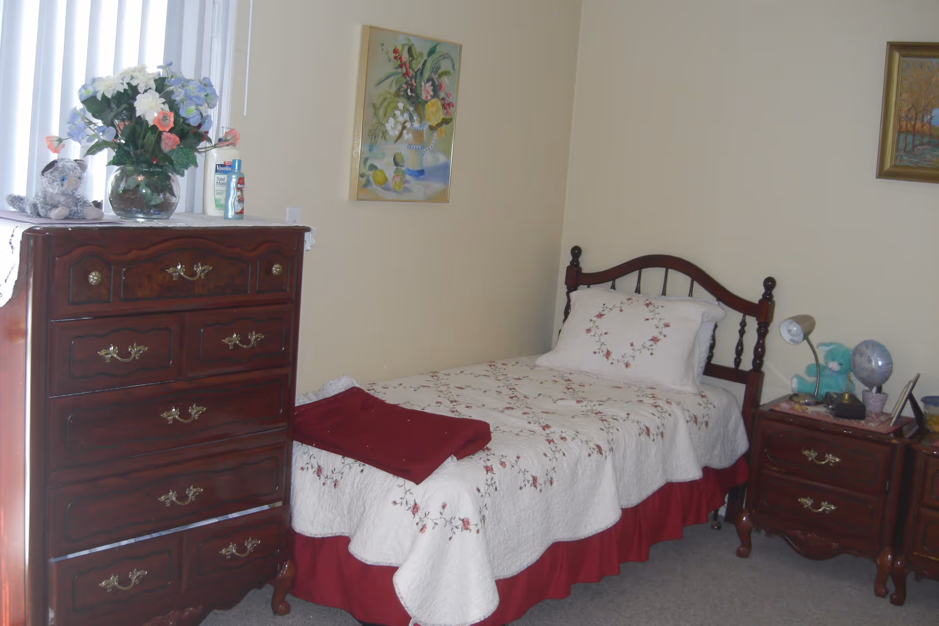 A bedroom with a single bed covered in a white quilt with floral embroidery and a red bed skirt. Next to the bed is a wooden nightstand with a lamp, a blue teddy bear, a globe, and other small items. On the left side of the bed is a tall wooden dresser with brass handles, topped with a vase of colorful flowers, a stuffed animal, and some lotion bottles. The walls are light-colored and decorated with two framed paintings.
