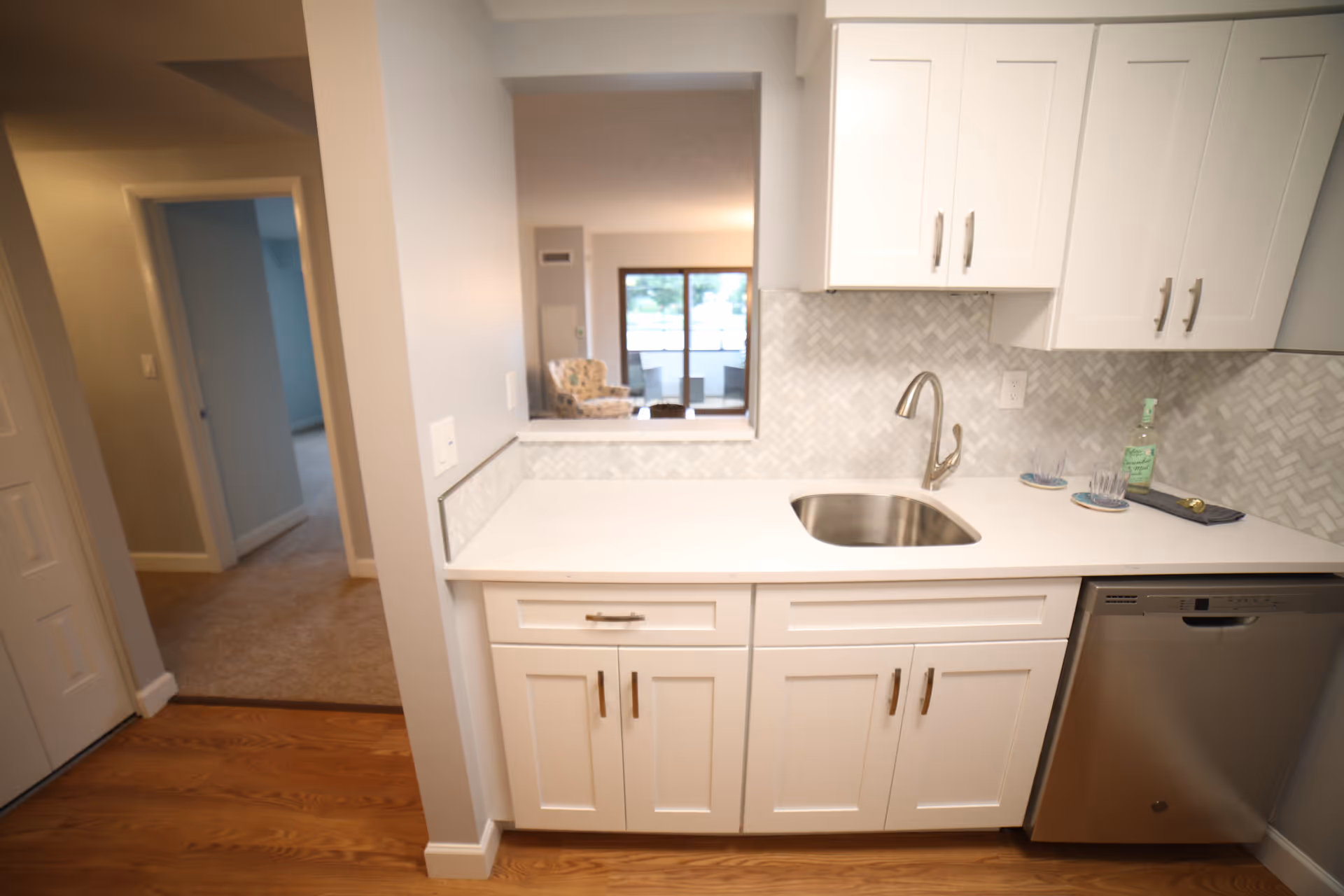 A modern kitchen area with white cabinets, a stainless steel sink, and a dishwasher. The countertop is white with a herringbone tile backsplash. There is a small pass-through window opening to a living room area with a chair and sliding glass doors leading outside. The floor is wood, and a hallway with carpet leads to other rooms.