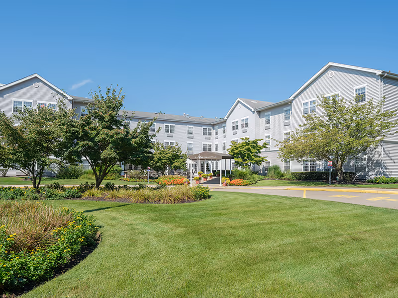 Exterior view of a multi-story senior living facility with green lawn, trees, and entrance canopy.