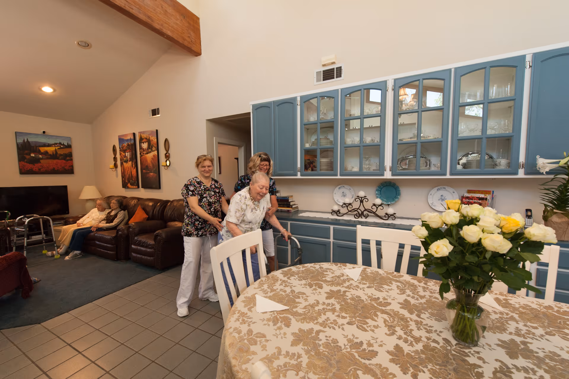 A senior living facility common area with a round dining table covered with a floral tablecloth and a vase of white and yellow roses. In the background, two elderly women are seated on a brown leather couch, while two caregivers assist an elderly woman using a walker near blue cabinets with glass doors displaying dishes and glassware. The room has a high ceiling with wooden beams and wall art.