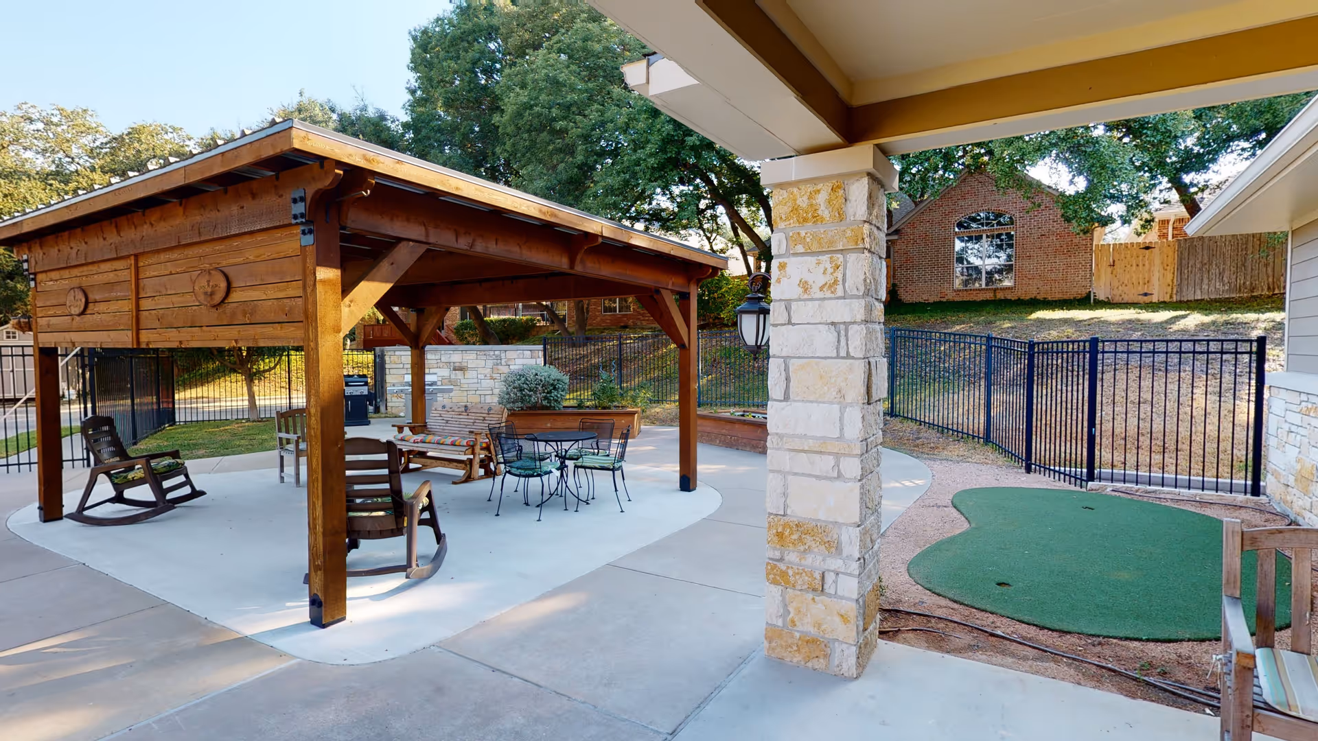 Outdoor patio area with a wooden pergola covering a seating area that includes rocking chairs and a metal table with chairs. There is a small putting green to the right, stone pillars, a black metal fence, and trees in the background.